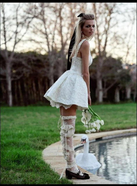Woman in white dress and lace stockings poses near pool with swan float. The shot is outdoors with soft lighting, trees in background.
