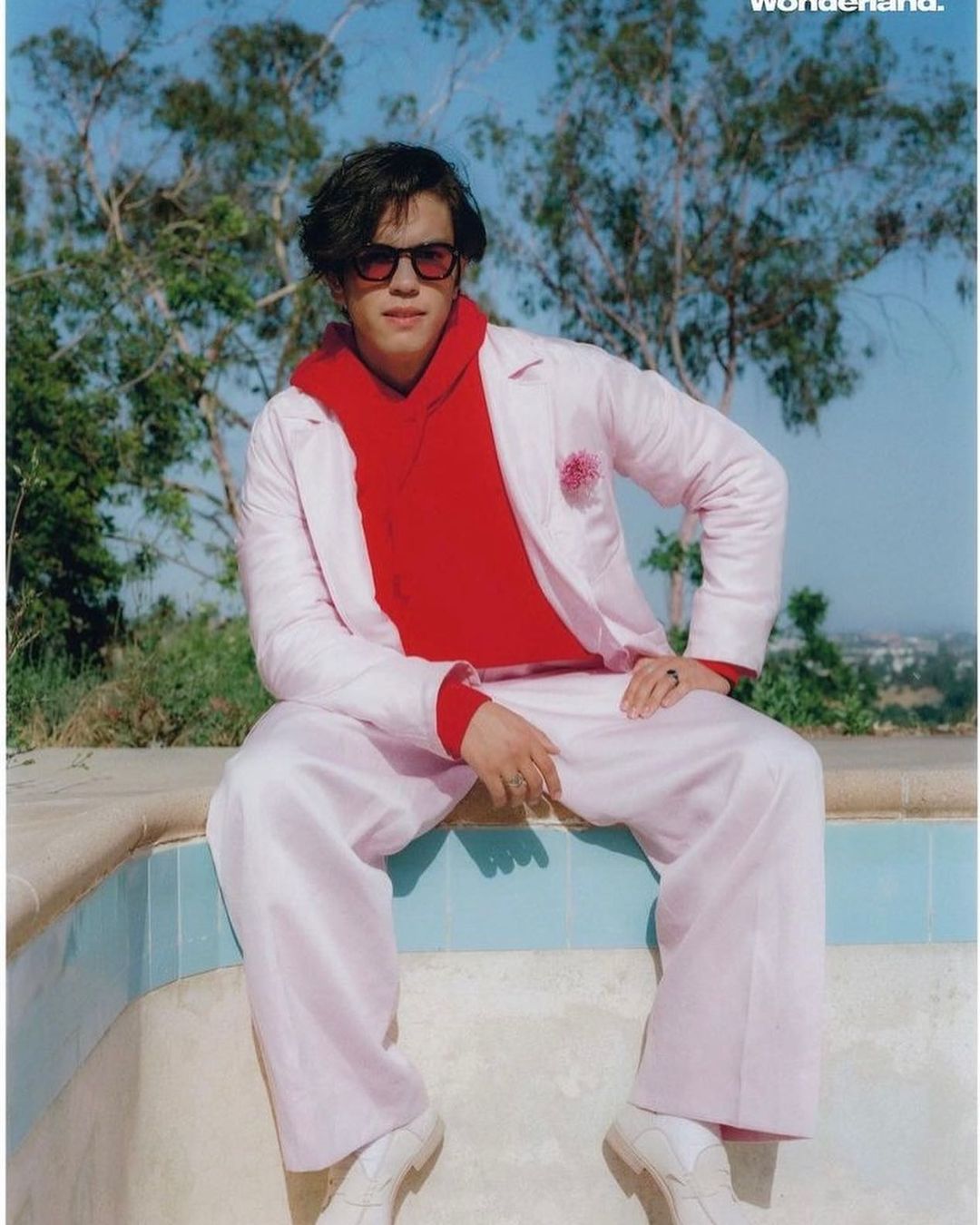 Man in pink suit and red hoodie sitting outside with trees and buildings in the background, wearing sunglasses, portrait orientation.