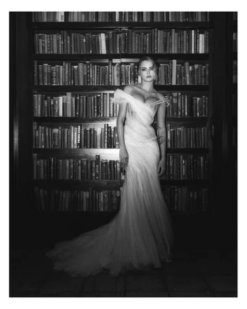 Woman in white off-shoulder gown standing in a library with rows of books in the background in a black and white portrait photograph.