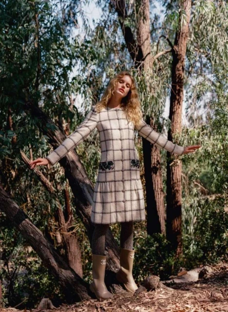 A woman in a patterned dress stands in a forest clearing with her arms outstretched on a sunny day, wearing beige boots with logos.