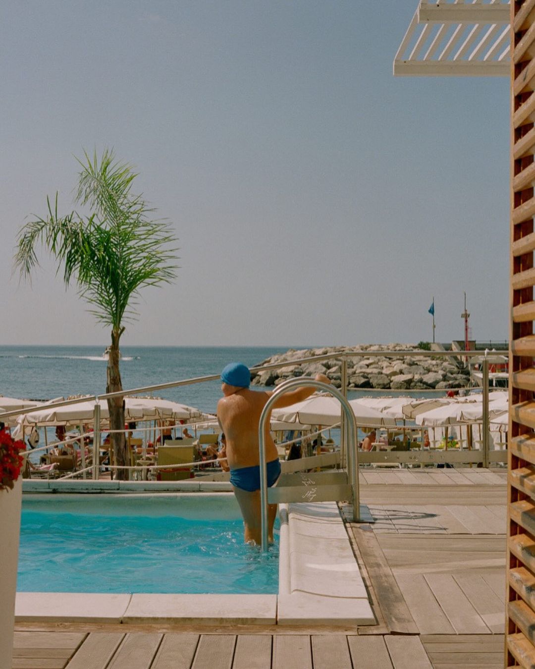 Person exiting outdoor pool next to palm tree, deck chairs, ocean, and coastline under a clear sky in bright daylight on a summer day.