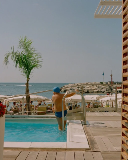 Person exiting outdoor pool next to palm tree, deck chairs, ocean, and coastline under a clear sky in bright daylight on a summer day.