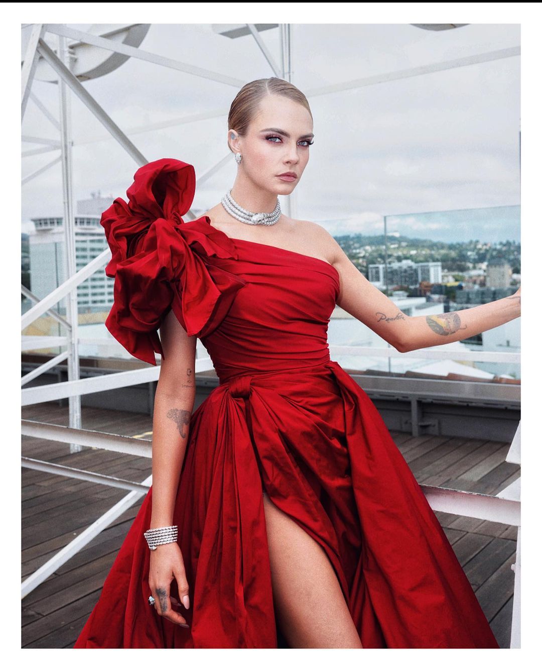 Cara Delevingne in a red, one-shoulder gown with a large bow detail, posing on a rooftop with a cityscape in the background.
