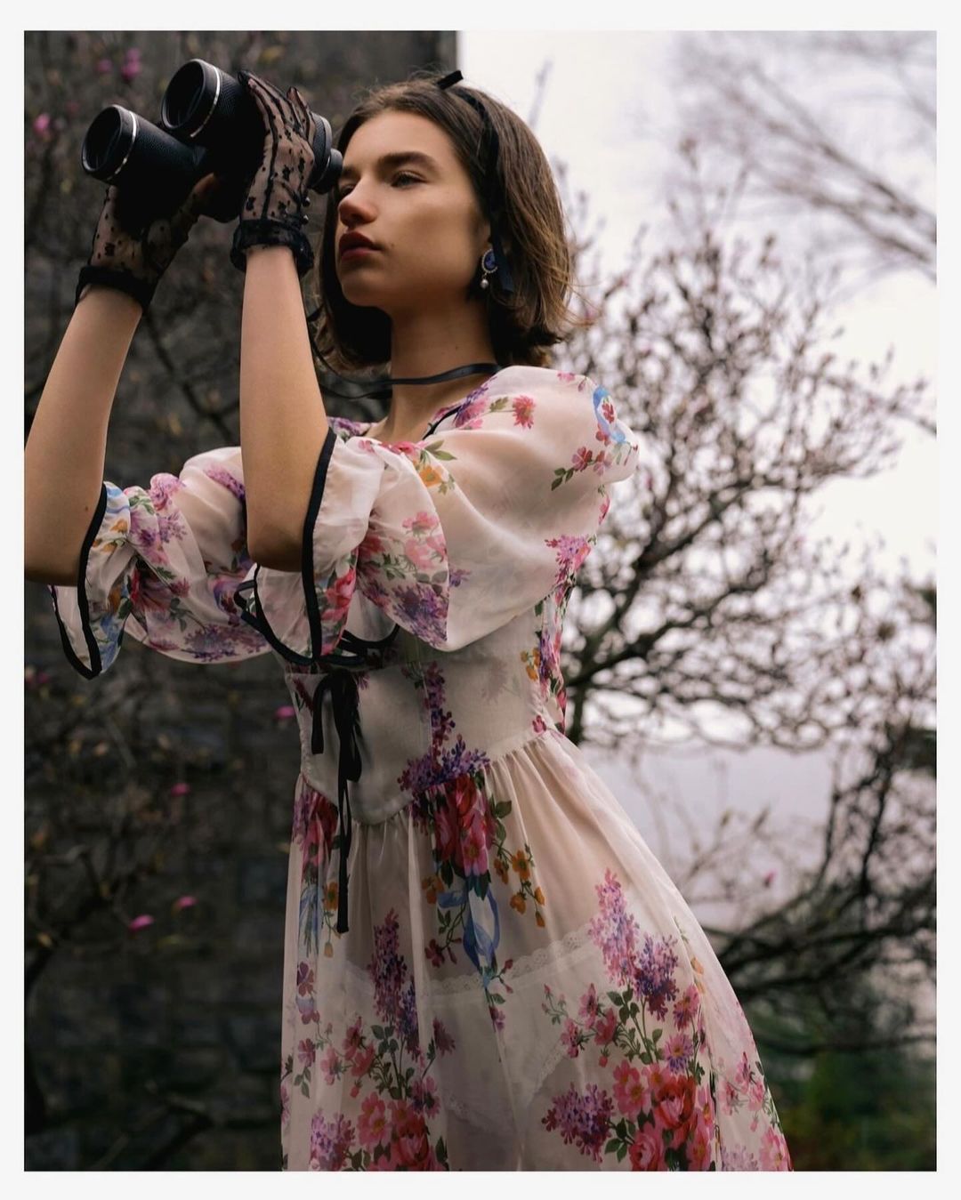Young woman in floral dress and lace gloves observes through binoculars in a natural outdoor setting with muted, dreamy lighting.