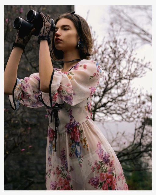 Young woman in floral dress and lace gloves observes through binoculars in a natural outdoor setting with muted, dreamy lighting.