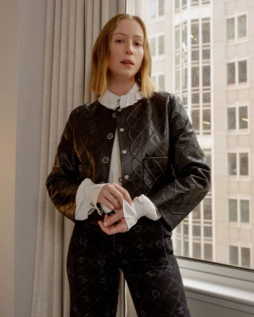 Indoor portrait of a woman wearing a black quilted jacket, white blouse, and black pants, standing in front of a large window.