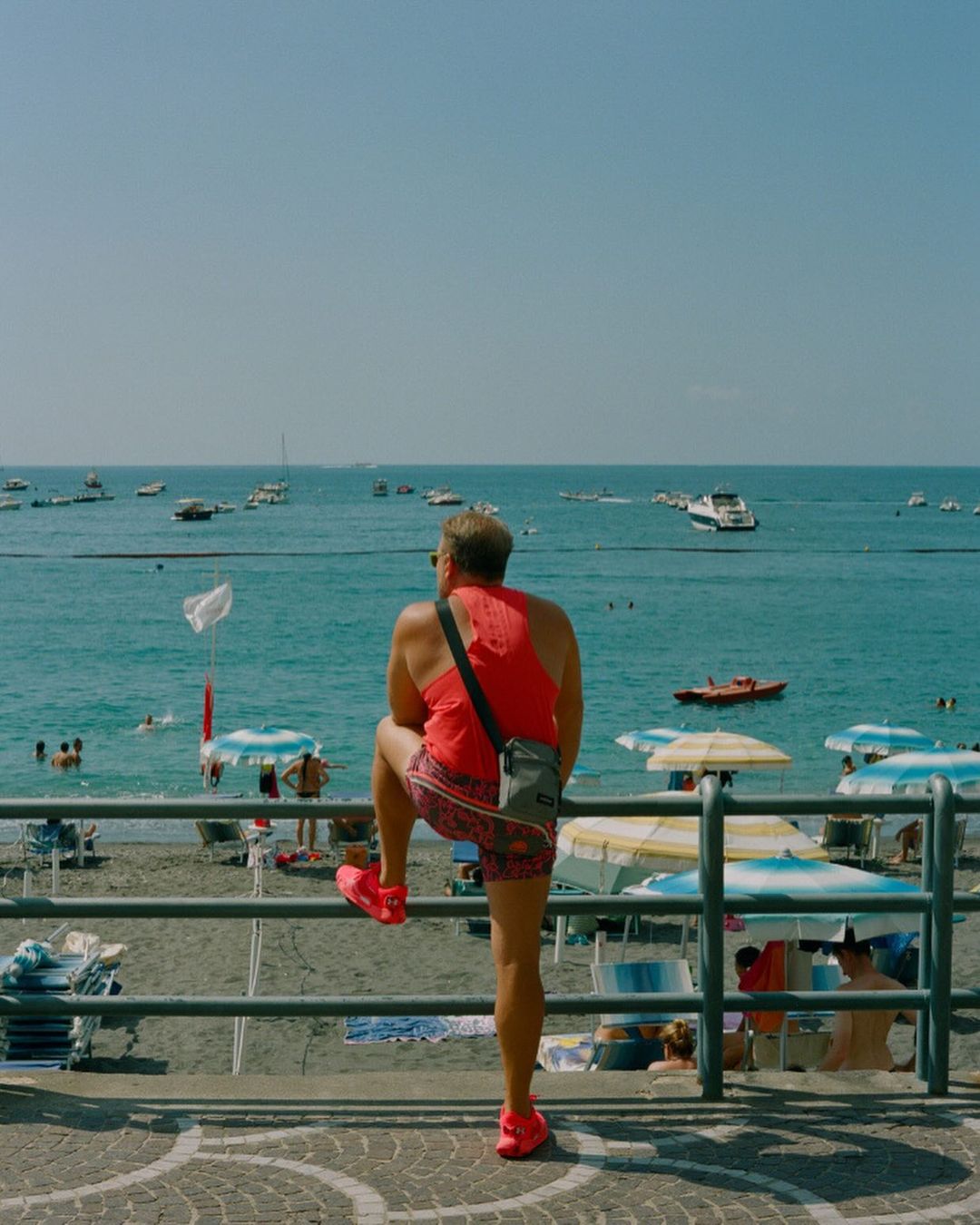 Man stands on beach railing overlooking ocean, wearing a red tank top and patterned shorts with boats and beachgoers in the background.