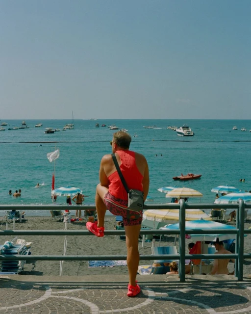 Man stands on beach railing overlooking ocean, wearing a red tank top and patterned shorts with boats and beachgoers in the background.