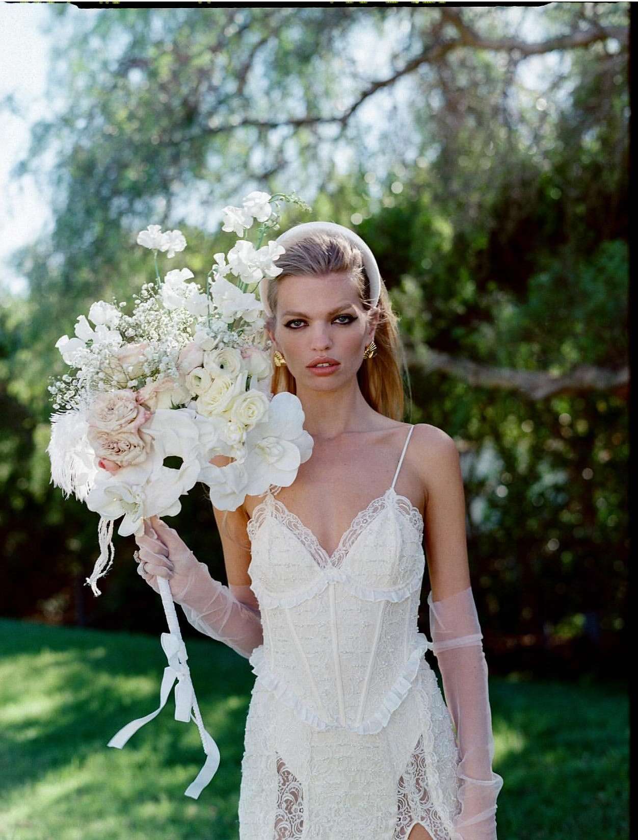 A blonde woman poses with a large bouquet of white flowers. She wears a white lace dress, sheer gloves, and a matching headband in a grassy outdoor setting.