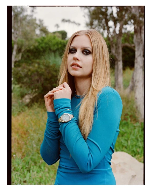 Blonde woman in turquoise dress poses outdoors. She has dramatic makeup, a wristwatch, and natural light illuminates the scene.
