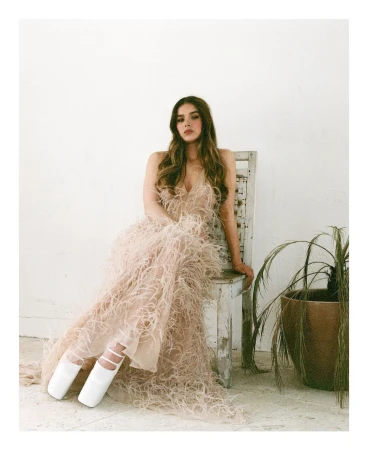 A woman with long brown hair sits on a distressed wooden chair wearing a feathered gown, posed against a white wall with a potted plant.