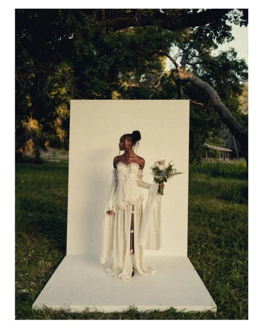An elegant Black woman poses outdoors against a white backdrop in a flowing gown, holding flowers, in a sunlit natural setting.