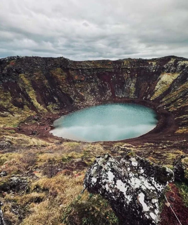 Crater lake landscape in Iceland with blue water, brown volcanic rock, and moody sky. Serene travel destination in nature.