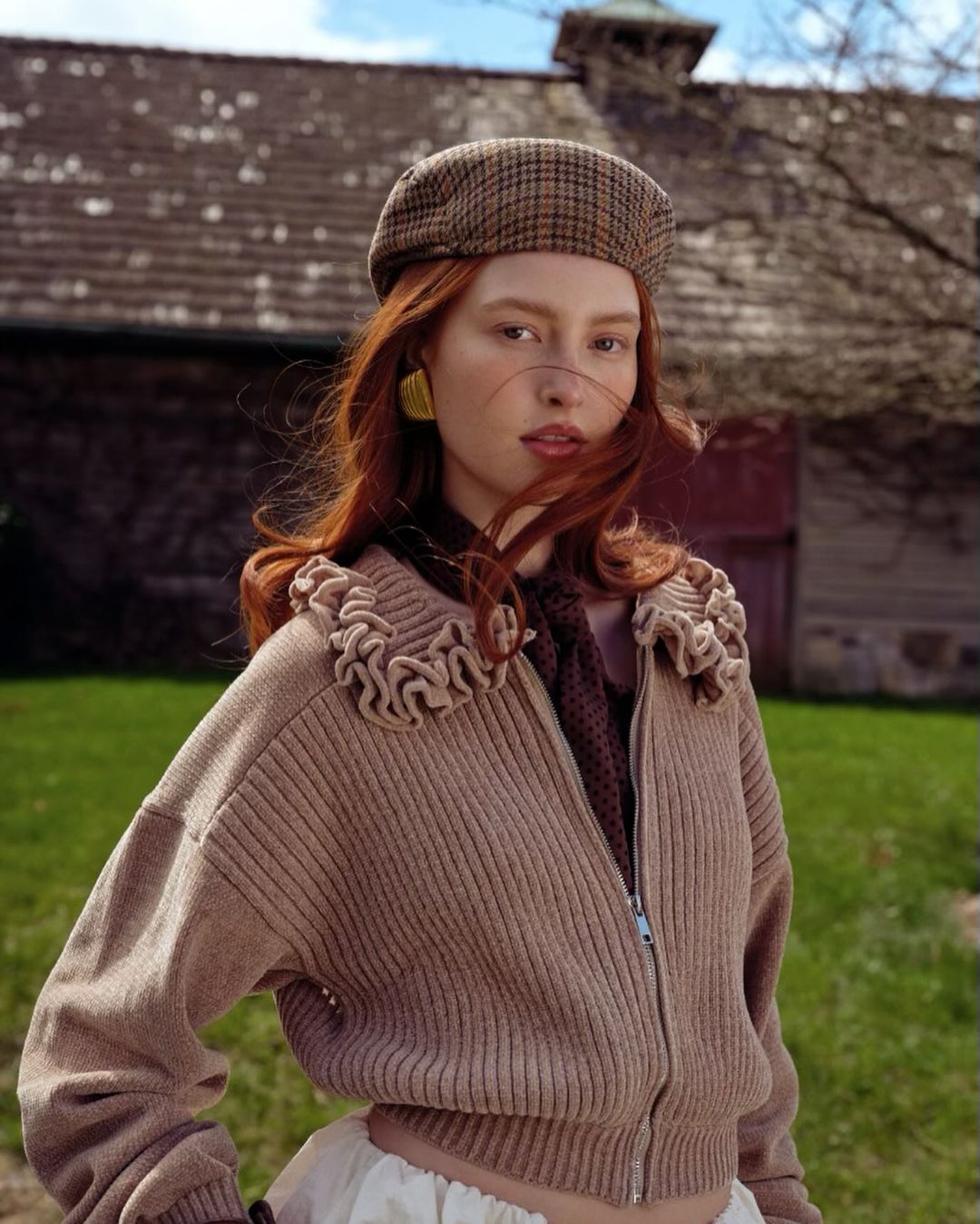 Portrait of a young woman with red hair wearing a brown plaid beret and a neutral-toned jacket with ruffled trim outdoors.