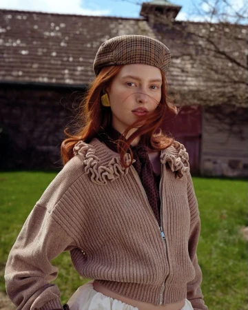 Portrait of a young woman with red hair wearing a brown plaid beret and a neutral-toned jacket with ruffled trim outdoors.