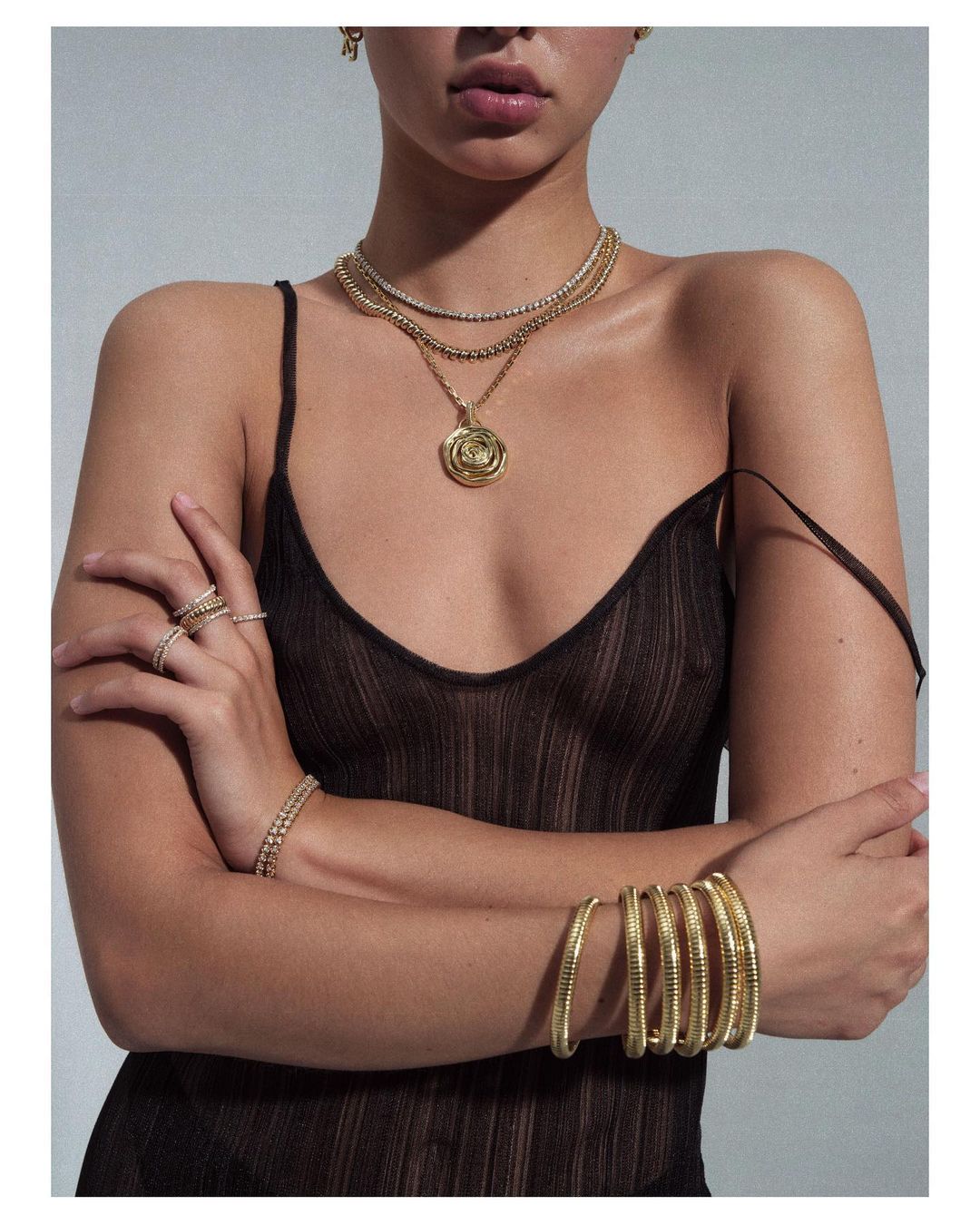 A woman with tan skin wearing jewelry and a see-through brown top poses against a grey background, showcasing fashion and beauty.