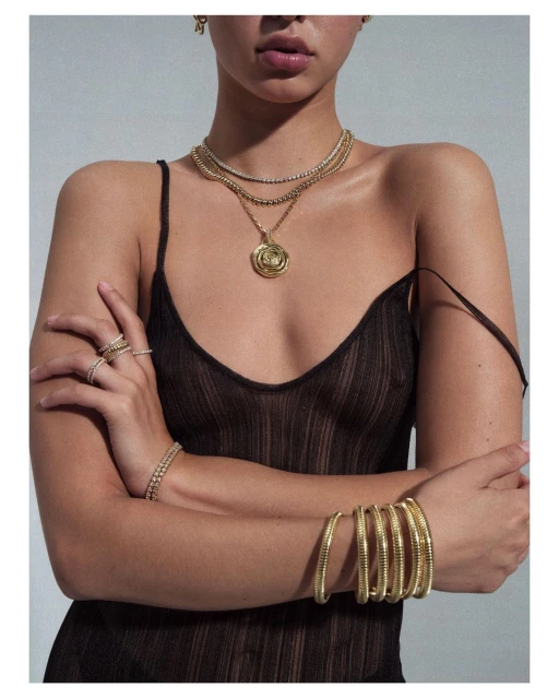 A woman with tan skin wearing jewelry and a see-through brown top poses against a grey background, showcasing fashion and beauty.