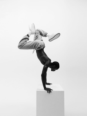 An adult male is doing a handstand on a white block wearing sweatpants and shoes in a studio with a white background in black and white.