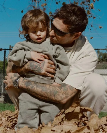 Father holds toddler outside in a pile of leaves on a sunny day. The man has tattoos covering both arms and is wearing sunglasses.