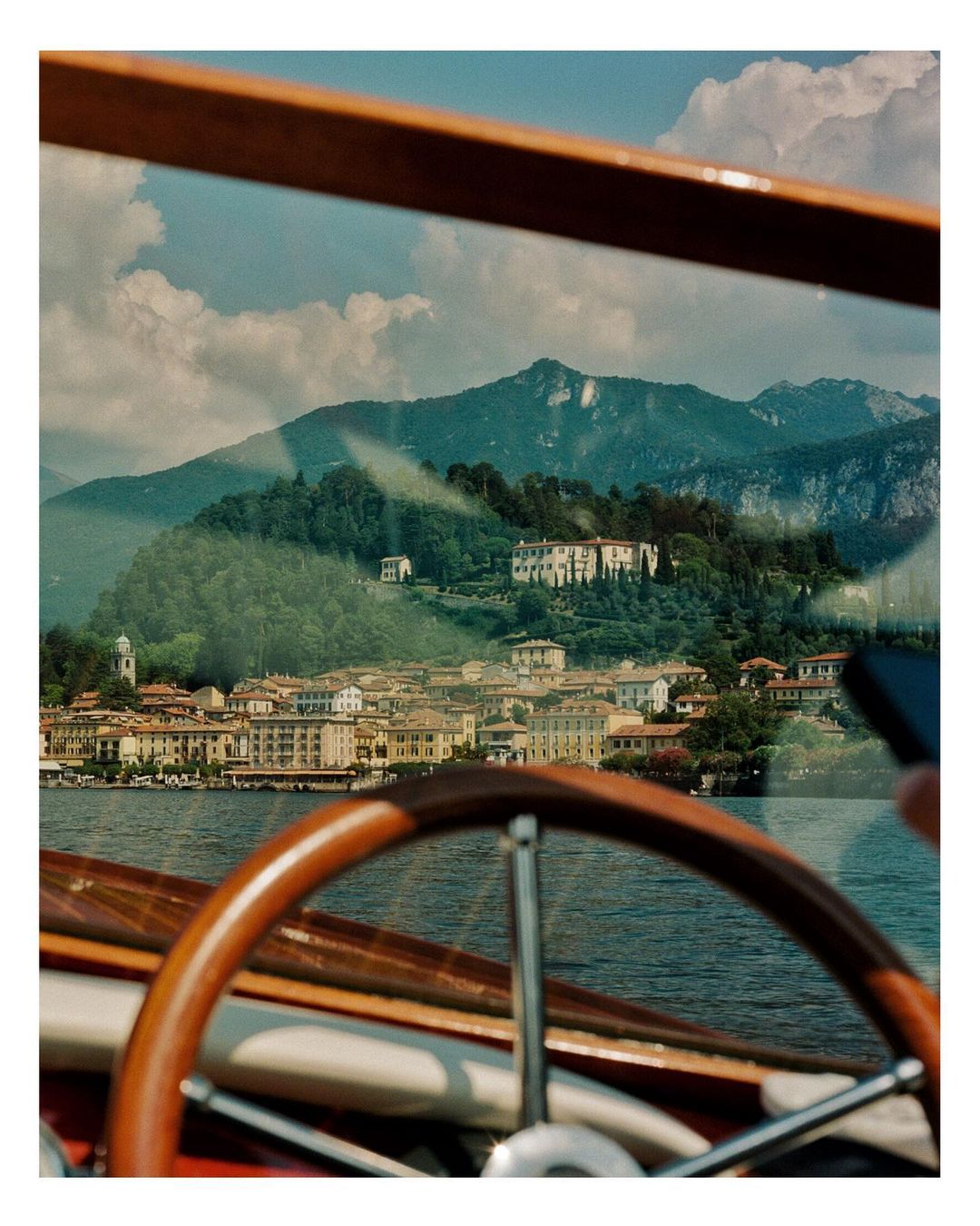 View from inside a wooden boat of buildings on Lake Como, Italy, with lush green mountains and a cloudy sky in the background.