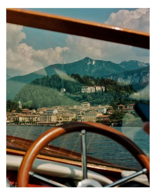 View from inside a wooden boat of buildings on Lake Como, Italy, with lush green mountains and a cloudy sky in the background.