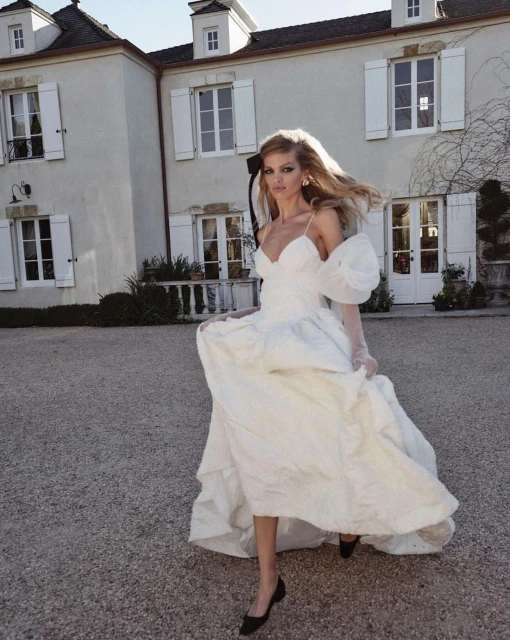 Woman in white formal gown with puffy sleeves walks on stone in front of French Provincial house with white shutters and dormer windows.