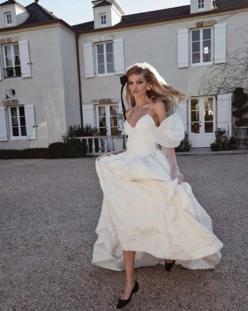 Woman in white formal gown with puffy sleeves walks on stone in front of French Provincial house with white shutters and dormer windows.