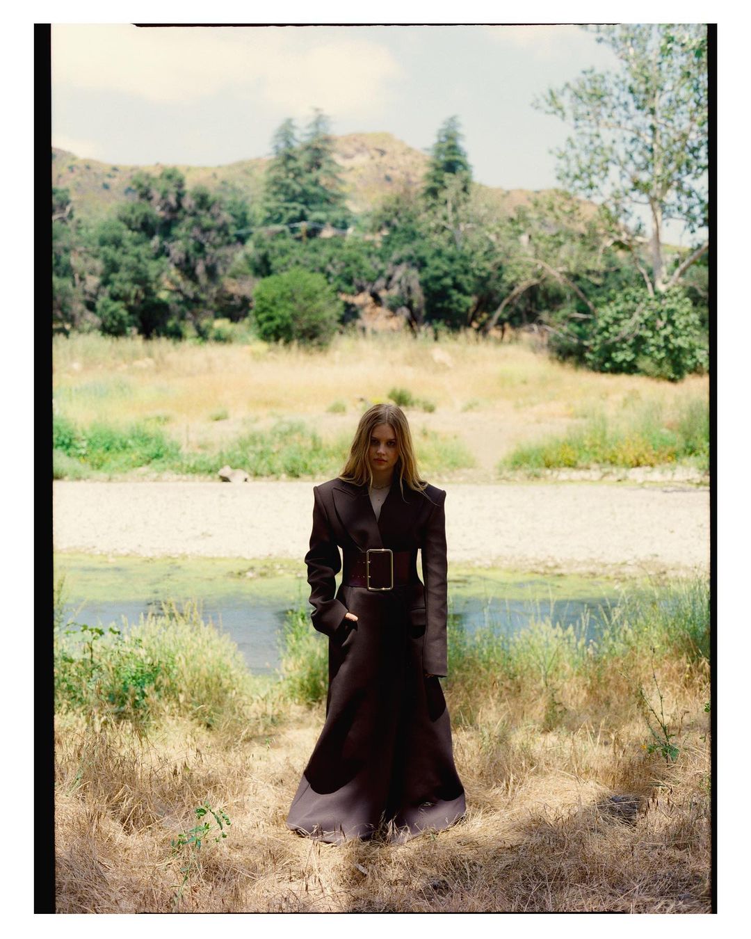 Model standing outdoors wearing a long dark belted coat against a backdrop of sparse trees and dry grass hills.