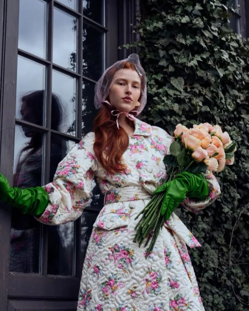 Red-haired woman in a floral coat holds peach roses outdoors. She wears green gloves and a netted headpiece with a door and ivy.