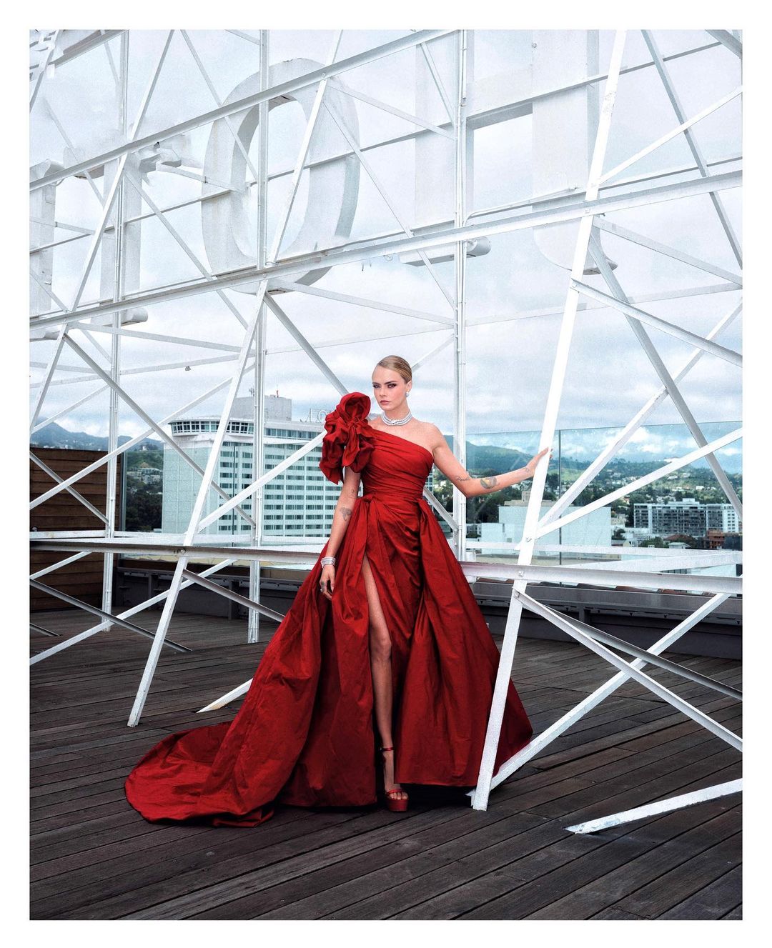 A woman in a red gown poses on a rooftop with a city view. The dress features a thigh-high slit and a large floral detail on the shoulder.