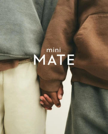 Close-up of two children holding hands, wearing hoodies and casual clothing, against a neutral background. Text "mini MATE" displayed.