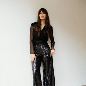 Brunette woman in black sheer top and sequin pants posing against a neutral wall in a studio, fashion portrait, waist up.