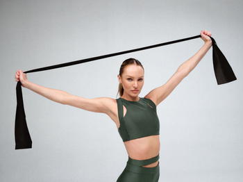 Athletic woman with long hair stretches a black resistance band in a studio setting with neutral background and soft lighting.