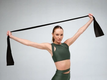 Athletic woman with long hair stretches a black resistance band in a studio setting with neutral background and soft lighting.
