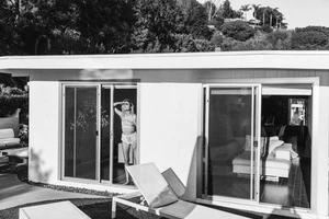 Black and white photograph of a woman in a doorway of a modern home, looking out toward the landscape with a suitcase.