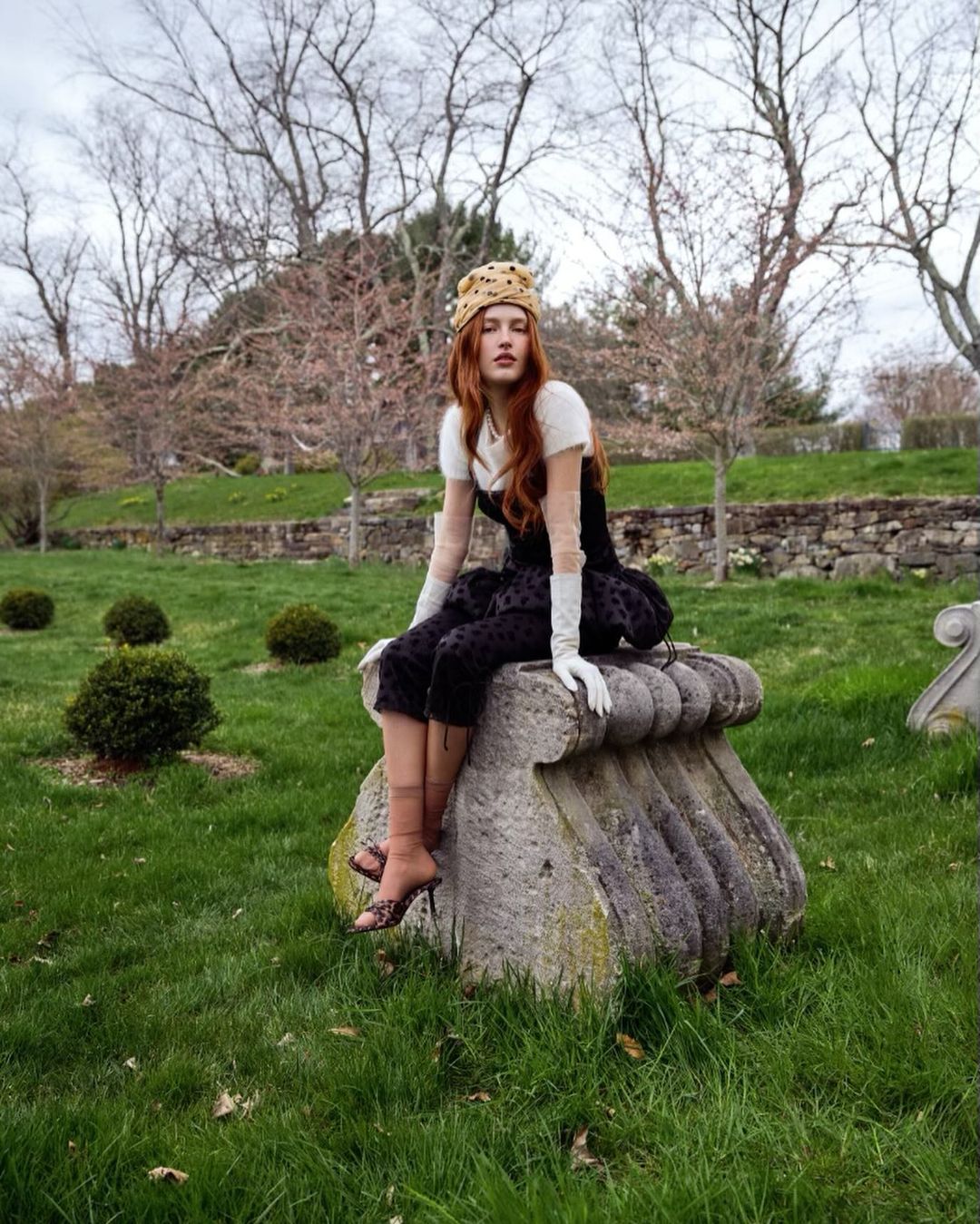 A red-haired woman in a textured hat and black dress poses on a stone pillar in an outdoor setting with trees and green grass.