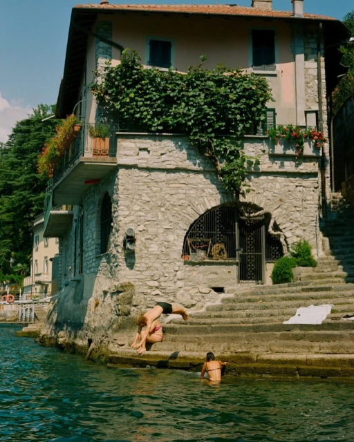 Two young people are swimming in a lake near a stone building with stairs. A third person sits on the steps near the water's edge.