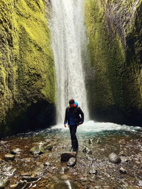A man stands in a shallow river in front of a cascading waterfall, surrounded by moss-covered cliffs and rocks under natural lighting.