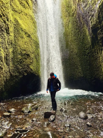 A man stands in a shallow river in front of a cascading waterfall, surrounded by moss-covered cliffs and rocks under natural lighting.