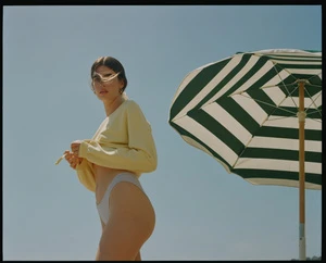 Adult woman poses near a striped parasol on a sunny day, wearing a yellow sweater and white bikini bottoms against a blue sky.