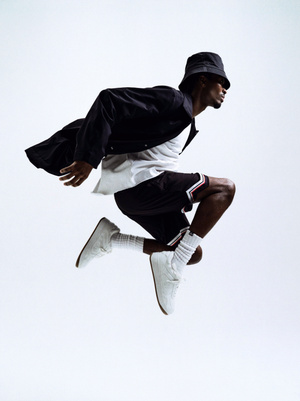 Black man mid-air jumping wearing a black bucket hat, dark jacket, white t-shirt, shorts, and white sneakers against a white background.