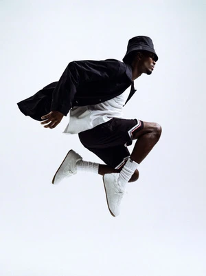 Black man mid-air jumping wearing a black bucket hat, dark jacket, white t-shirt, shorts, and white sneakers against a white background.