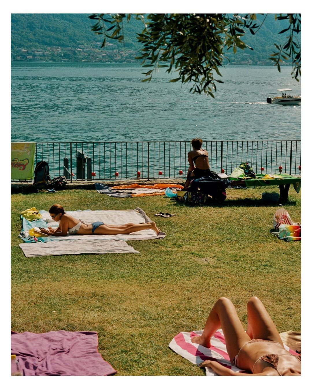 Three adult women relax on towels on a grassy area with water in the background, under a sunny sky during daylight hours.