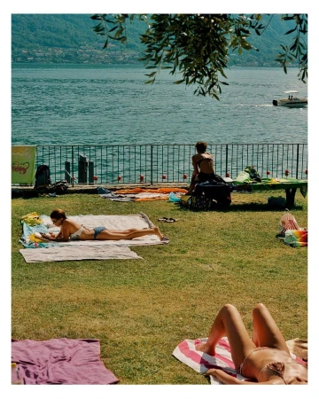 Three adult women relax on towels on a grassy area with water in the background, under a sunny sky during daylight hours.