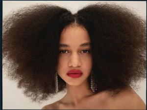 Close-up portrait of a young adult woman with large afro, wearing red lipstick and dangling crystal earrings against a white background.