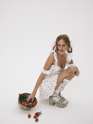 Woman in floral dress crouching with strawberries in basket in white studio, wearing platform shoes with ribbons in her blonde hair.