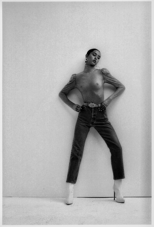 Model in sheer long sleeve top and high waisted jeans posing against a white studio wall in black and white