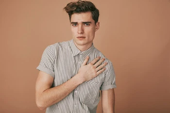 Young man wearing a grey striped short-sleeve button-down shirt with his hand on his chest against a warm beige background.