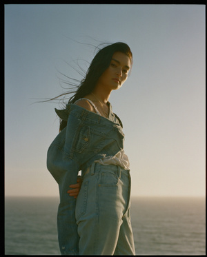 Young woman in denim jacket stands on a coastline with the ocean and sky visible in the background in a natural light photograph.