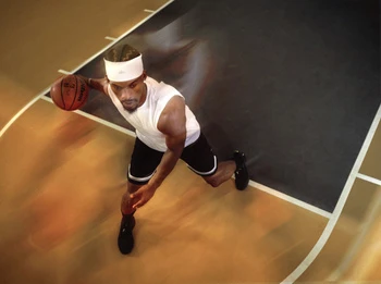 Overhead view of an African American male basketball player dribbling on an indoor court, wearing a white headband, tank top, and black shorts.
